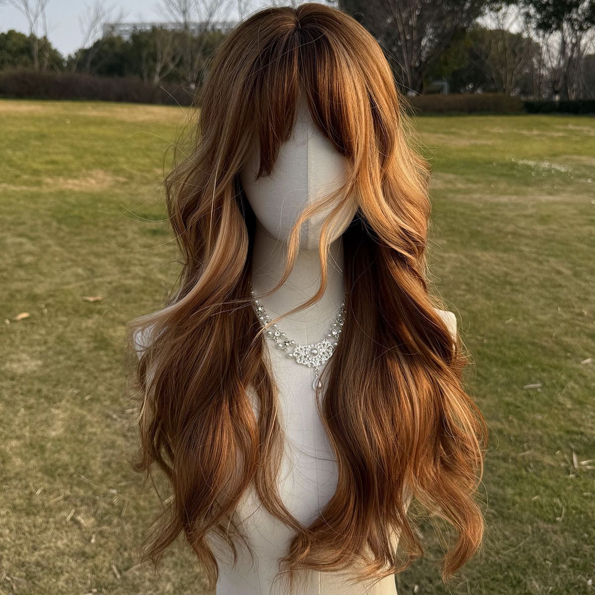 Long auburn-brown wavy wig with bangs and lighter highlights, draped over shoulders.